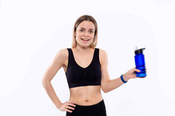 Joven mujer rubia deportiva en una ropa deportiva negra con relojes inteligentes sostiene la botella con agua después del entrenamiento de pie sobre fondo blanco
. - Foto, imagen