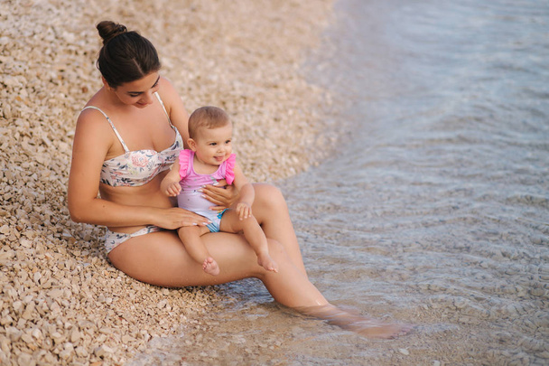 junge Mutter spielt mit ihrem kleinen Mädchen am Strand am Meer. Mutter und Tochter im Badeanzug - Foto, Bild