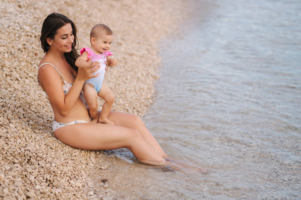 Beautiful young mother with her cute baby girl enjoying summer sun on the beach. Happy girls in swimsuit by the sea. Baby laughing - Photo, Image