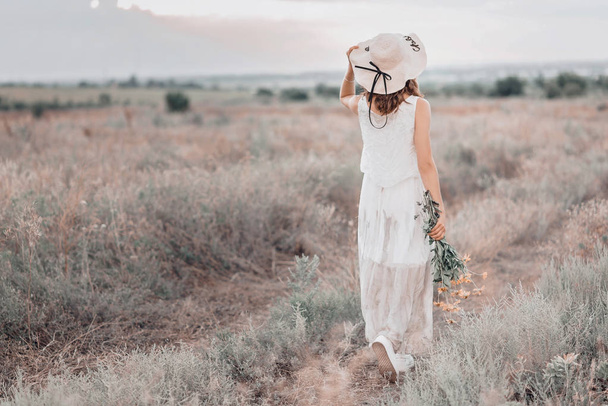 Mujer joven en sombrero de paja y ramo de flores caminando en el campo de verano
. - Foto, Imagen