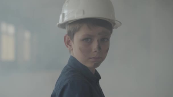 Portrait adorable boy in a protective helmet turning and looking at the camera in the background of smoke indoors. Concept of fire, disaster, flammability, non-compliance with safety rules. - Πλάνα, βίντεο