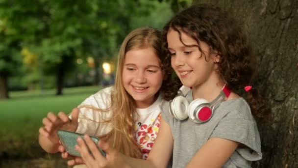 Δύο χαριτωμένες νεαρές αδελφές με ακουστικά που διασκεδάζουν στην όμορφη μέρα. Χαρούμενα παιδιά παίζουν στο καλοκαιρινό πάρκο. Τα παιδιά παρακολουθούν κάτι ενδιαφέρον στο τηλέφωνο. Καλοκαιρινές δραστηριότητες για παιδιά. - Πλάνα, βίντεο
