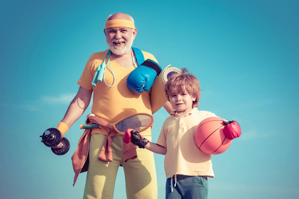 Senior man and child exercising on blue sky. Sport exercise for kids. Portrait of a healthy father and son working out over blue sky background. - Photo, Image