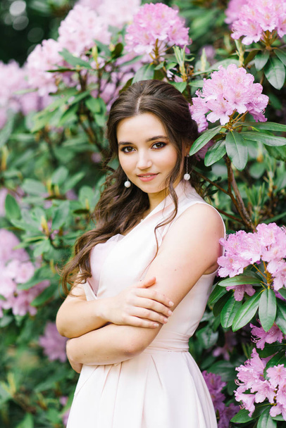 Beautiful portrait of a lovely girl who stands surrounded by lilac pink flowers blooming rhododendron in the spring in the garden. Beautiful clean skin of a young woman. Professional makeup - Fotografie, Obrázek
