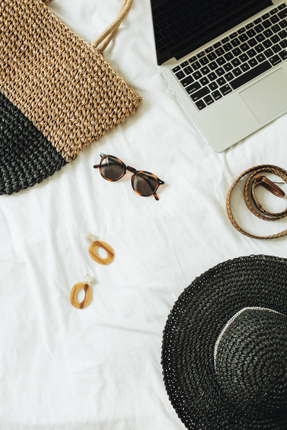 女性のファッションアクセサリー:メガネ、イヤリング、ベルト、麦わら帽子、わら袋、ノートパソコンは白いシートでベッドに横たわっています。夏と旅行のコンセプト。トップビュー、フラットレイ.  - 写真・画像