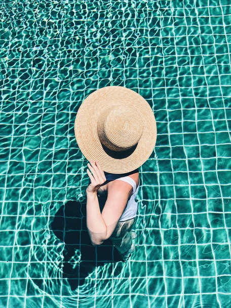 Yaz seyahat moda ve tatil konsepti. Hasır şapkalı ve beyaz mayosu olan genç ve tabaklanmış güzel bir kadın yüzme havuzunda duruyor. Üst görünüm, düz yatıyordu. - Fotoğraf, Görsel
