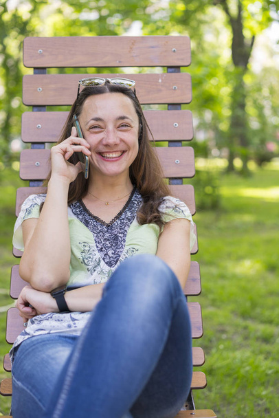 Mujer joven hablando en el teléfono inteligente y riendo en el parque en el banco Hermosa mujer relajándose en un banco del parque y utilizando un teléfono inteligente. tonificado
 - Foto, Imagen