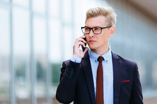 Young handsome Caucasian businessman in glasses talking on mobile phone in front of office building. Male executive manager in trendy clothes using smartphone outdoors. Modern technology concept - Foto, immagini