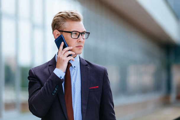 Young handsome Caucasian businessman in glasses talking on mobile phone in front of office building. Male executive manager in trendy clothes using smartphone outdoors. Modern technology concept - Photo, image