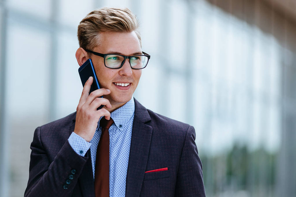 Young handsome Caucasian businessman in glasses talking on mobile phone in front of office building. Male executive manager in trendy clothes using smartphone outdoors. Modern technology concept - Photo, image