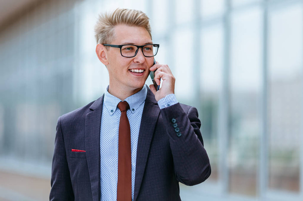 Young handsome Caucasian businessman in glasses talking on mobile phone in front of office building. Male executive manager in trendy clothes using smartphone outdoors. Modern technology concept - Valokuva, kuva
