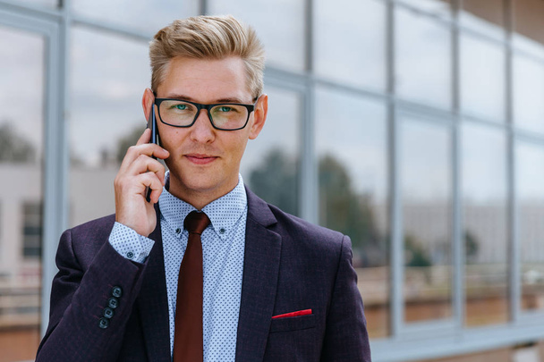 Young handsome Caucasian businessman in glasses talking on mobile phone in front of office building. Male executive manager in trendy clothes using smartphone outdoors. Modern technology concept - Φωτογραφία, εικόνα