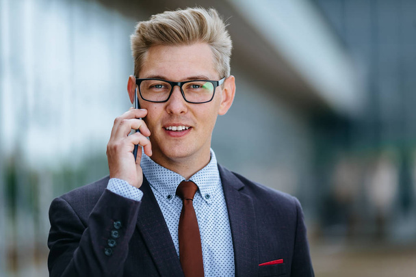 Young handsome Caucasian businessman in glasses talking on mobile phone in front of office building. Male executive manager in trendy clothes using smartphone outdoors. Modern technology concept - Fotoğraf, Görsel