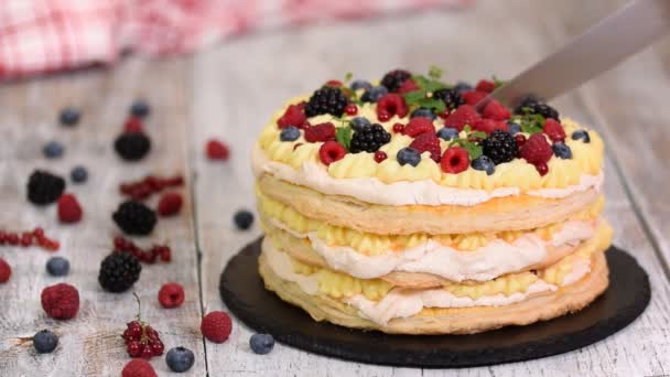 Layered cake decorated with berries and mint on table. Woman cutting delicious puff pastry cake. - Video