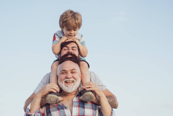 Ευτυχισμένη μέρα των πατέρων. Πατέρας και γιος απολαμβάνουν έξω. Ανδρική γενιά: ο παππούς πατέρας και ο εγγονός αγκαλιάζονται κοιτάζοντας την κάμερα και χαμογελούν. - Φωτογραφία, εικόνα
