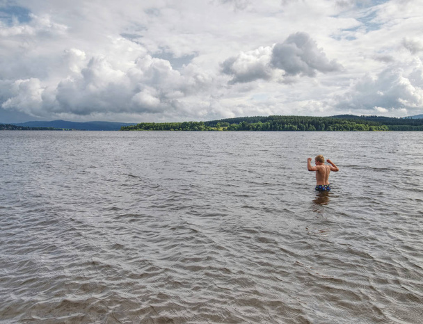 Niño en pantalones cortos de natación azul permanecer en el agua fría del lago
 - Foto, imagen