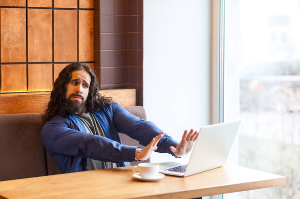 Angry handsome young adult man freelancer in casual style sitting in cafe and showing stop hands gesture to laptop  - Foto, Imagen