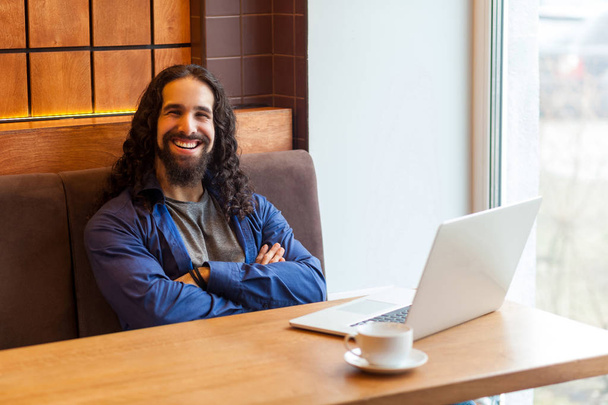Satisfied handsome bearded young man freelancer in casual style with crossing arms sitting with laptop at table and looking at camera with toothy smile in cafe - Photo, image