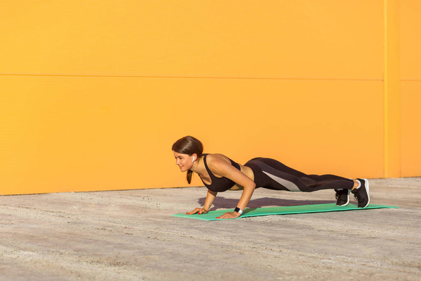 side view of muscular sporty strong young athletic woman in black sportswear standing in plank position on green mat in sunshine against orange wall background, chaturanga dandasana yoga posture - Fotó, kép