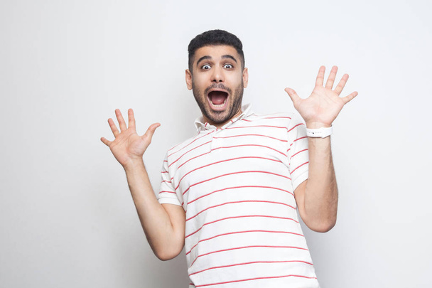 Portrait of surprised handsome bearded young man in striped t-shirt standing with raised arms and looking at camera with amazed shocked face isolated on white background - Фото, изображение