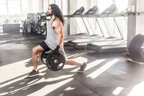 Side view of concentrated young adult sportsman athlete with long curly hair working out in gym while squatting on one knee and holding two disks, doing exercises for legs  - Φωτογραφία, εικόνα