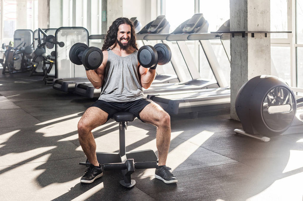grimace young adult muscular built athlete with long curly hair working out in gym while sitting on weightlifting machine and  holding two dumbbells with raised arms, exercises for swing shoulders  - Photo, image