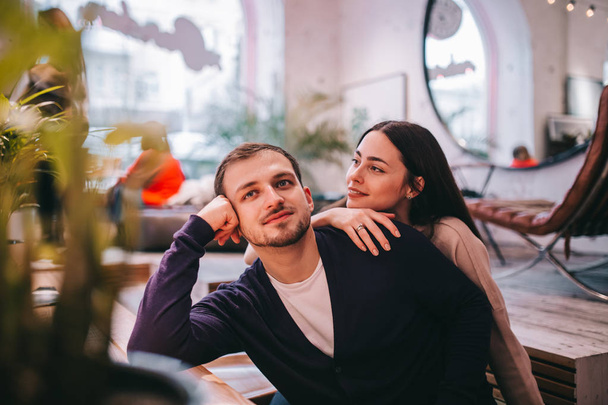 Loving girl holds her hand on the shoulder of her boyfriend sitting at the table in the cozy romantic cafe - Foto, Imagen