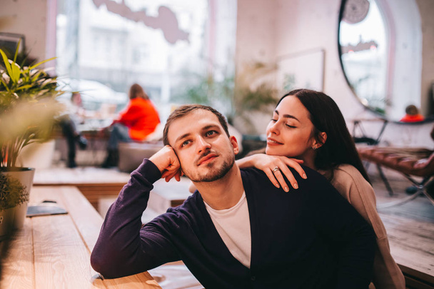 Loving girl holds her hand on the shoulder of her boyfriend sitting at the table in the cozy romantic cafe - Foto, imagen