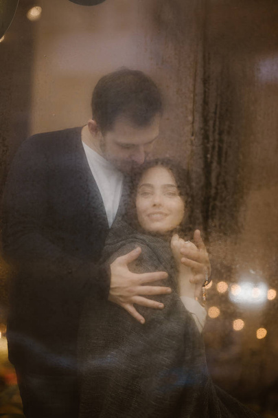 Romantic loving guy hugs his happy girlfriend standing behind a wet window with lights. - Photo, Image