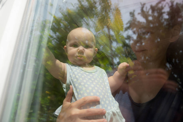 Το παιδί κοιτάζει έξω από το παράθυρο, το κοριτσάκι κοιτάζει έξω από το παράθυρο, και η μητέρα της την κρατά. - Φωτογραφία, εικόνα
