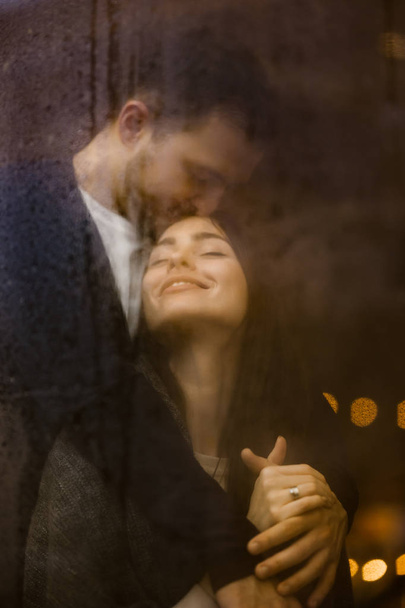 Loving guy hugs and kisses his happy girlfriend standing behind a wet window with lights. Romantic couple - Photo, image