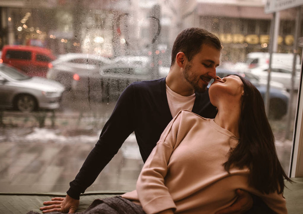 Romantic couple. Loving guy kisses his beautiful girlfriend sitting on the windowsill in a cozy cafe. - 写真・画像