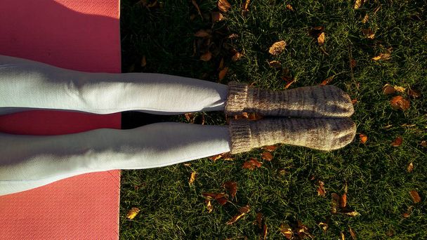 Yoga girl in autumn park with yoga mat on green carpet with fallen yellow leaves. Funny autumn legs wearing woolen socks and doing yoga in cold autumn park in the morning - Photo, image