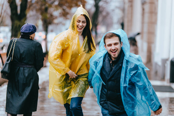 Funny and loving guy and girl in yellow and blue raincoats are running in the rain outside. - Фото, изображение