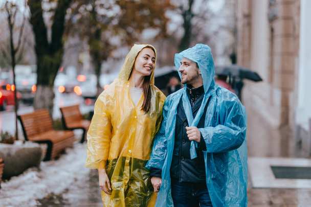 Loving guy and girl in the yellow and blue  raincoats walk on the street in the rain - Zdjęcie, obraz