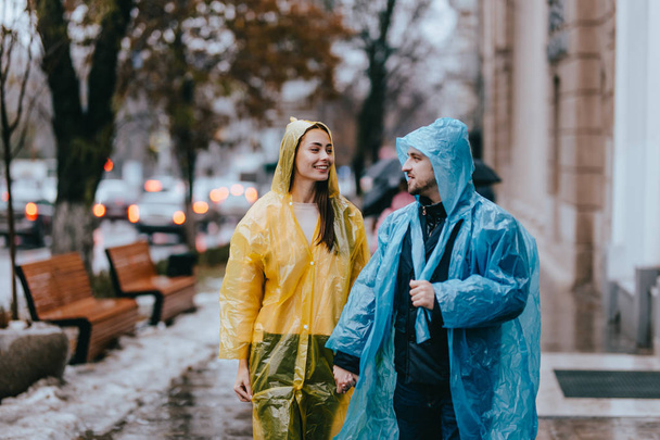 Loving guy and girl in the yellow and blue  raincoats stand on the street in the rain - Фото, зображення