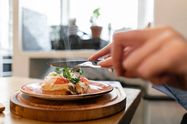 Cialda tradizionale belga con salmone, foglie di lattuga e uova in camicia. Una giovane donna sta facendo colazione in un caffè, mani con una forchetta e un coltello primo piano
. - Foto, immagini