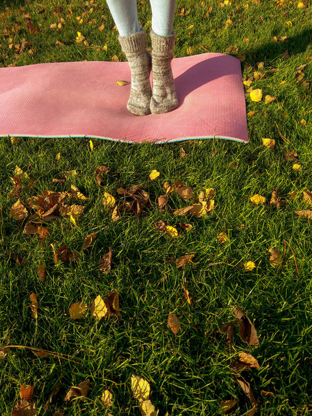 Yoga girl in autumn park with yoga mat on green carpet with fallen yellow leaves. Funny autumn legs wearing woolen socks and doing yoga in cold autumn park in the morning - Photo, Image