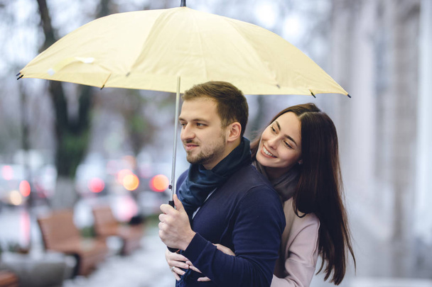 Χαρούμενο ρομαντικό ζευγάρι, ο τύπος και η κοπέλα του ντυμένοι με casual ρούχα αγκαλιάζονται κάτω από την ομπρέλα και κοίτα ο ένας τον άλλο στο δρόμο στη βροχή. - Φωτογραφία, εικόνα
