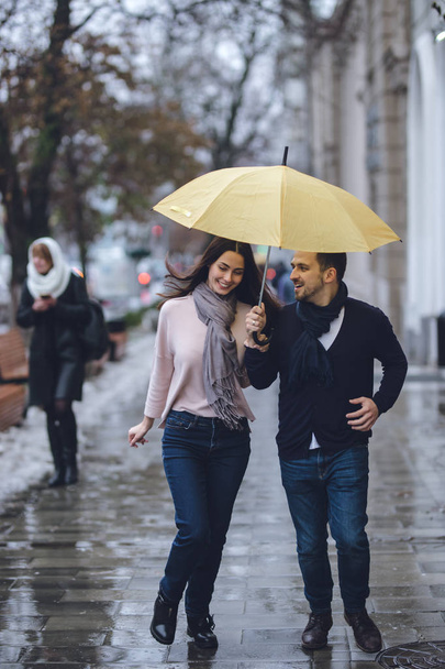 Beautiful couple, guy and his girlfriend dressed in casual clothes are running under the umbrella on the street in the rain. - Фото, изображение
