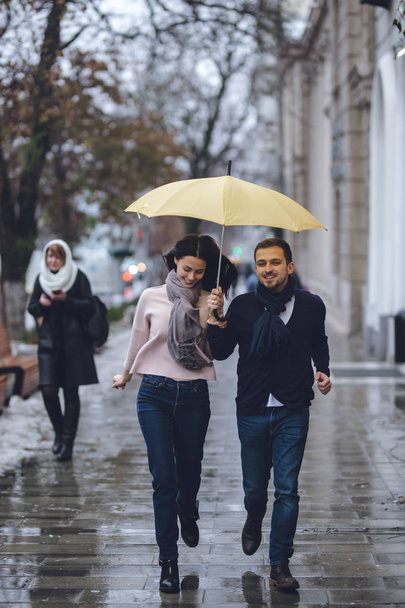 Beautiful couple, guy and his girlfriend dressed in casual clothes are running under the umbrella on the street in the rain. - Photo, Image
