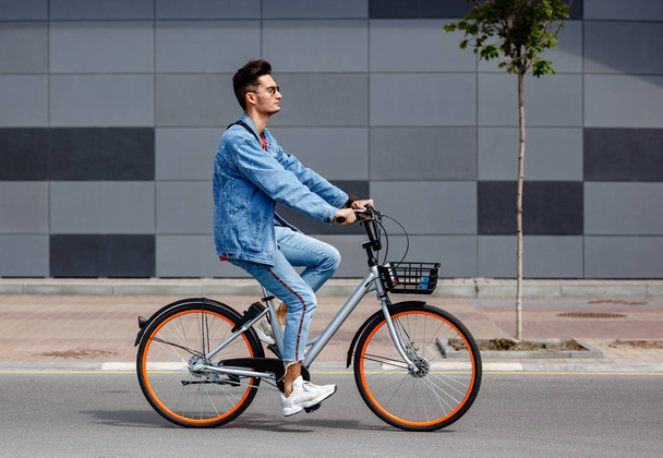 Şık adam moda kot giysiler giymiş gri bir binanın arka planına karşı sokakta bisiklet sürmek - Fotoğraf, Görsel
