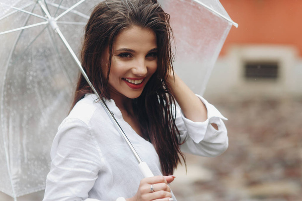 Νεαρό κομψό κορίτσι που ποζάρει στο δρόμο της πόλης, με τα πόδια γύρω από την πόλη με ομπρέλα την καλοκαιρινή μέρα. Είναι καλοκαιρινή βροχή.. - Φωτογραφία, εικόνα