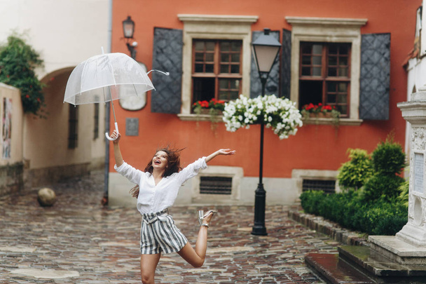 Ευτυχισμένη επιτυχημένη γυναίκα τουρίστρια περπατώντας γύρω από το αστικό σκηνικό, παίζοντας με την ομπρέλα της. Είναι καλοκαιρινή βροχή.. - Φωτογραφία, εικόνα