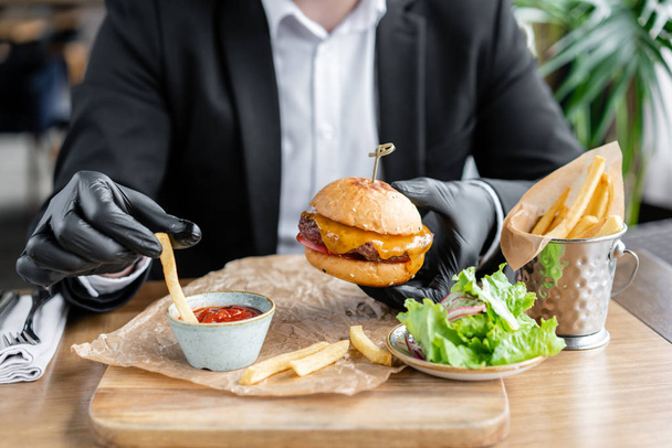 Young man in business suit and black gloves eating delicious juicy cheeseburger with beef chop. Burgers in the cafe with salad and fries, fast food. - Fotó, kép