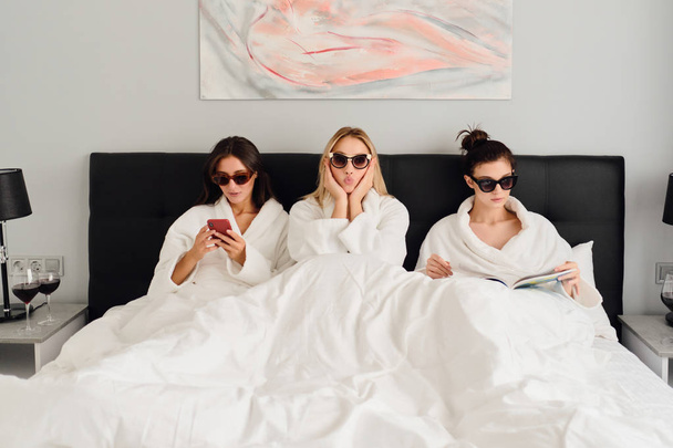 Three young beautiful women in white bathrobes and sunglasses lying in bed in modern cozy hotel. Pretty girl dreamily looking in camera while two girls near using cellphone and reading book - Fotoğraf, Görsel