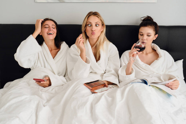 Three pretty women in white bathrobes lying in bed in modern cozy hotel. Cute girl making faces holding eye shadow palette in hands while girls near happily using cellphone and reading book with wine - Photo, Image