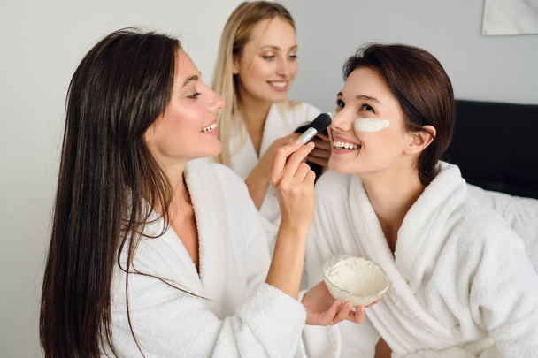 Three young beautiful women in white bathrobes sitting on bed happily making cosmetic mask together in modern cozy hotel  - 写真・画像