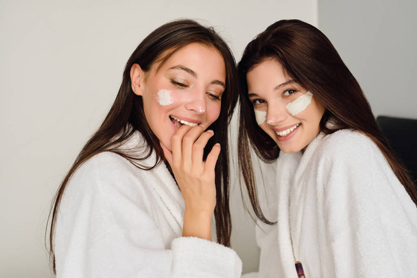 Két fiatal, gyönyörű nők Sötét haja fehér fürdőköpenyben ül az ágyban, kozmetikai maszk arcok boldogan keres a kamera modern szálloda  - Fotó, kép