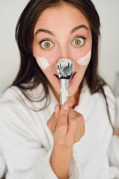 Jovem mulher muito alegre em roupão de banho branco com máscara cosmética no rosto segurando escova perto do nariz olhando feliz na câmera no moderno hotel acolhedor
  - Foto, Imagem